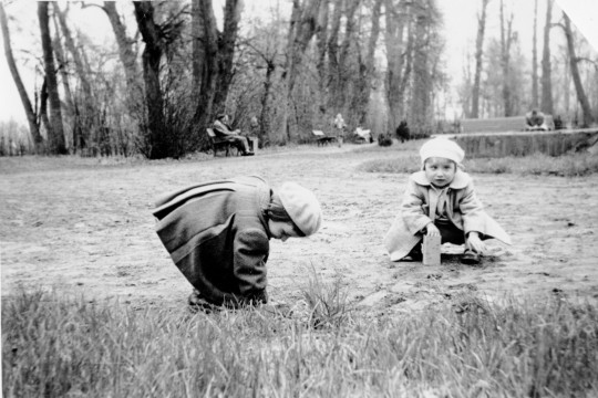 Park, ujęcie z poziomu dziecka. Dwoje dzieci kuca na trawie. Ubrane w berety i płaszczyki. W tle ludzie siedzą na ławkach albo spacerują.