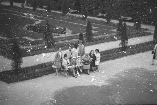 Fotografia lekko zniszczona, pełna małych białych plamek. Widok z Tarasu Górnego na grupę osób. Niektórzy siedzą na ławce, cztery osoby stoją za nią.