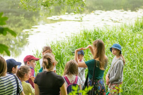 Grupa dzieci nad stawem, które stoją w trawie. Młoda kobieta pokazuje im coś rękami. Dzieci patrzą na to co robi.