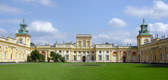 Kolorowe zdjęcie współczesne. Przedstawia widok na Pałac Wilanowski w słońcu. Przed pałacem płat zielonej równej trawy. Nad pałacem błękitne niebo z chmurami. 