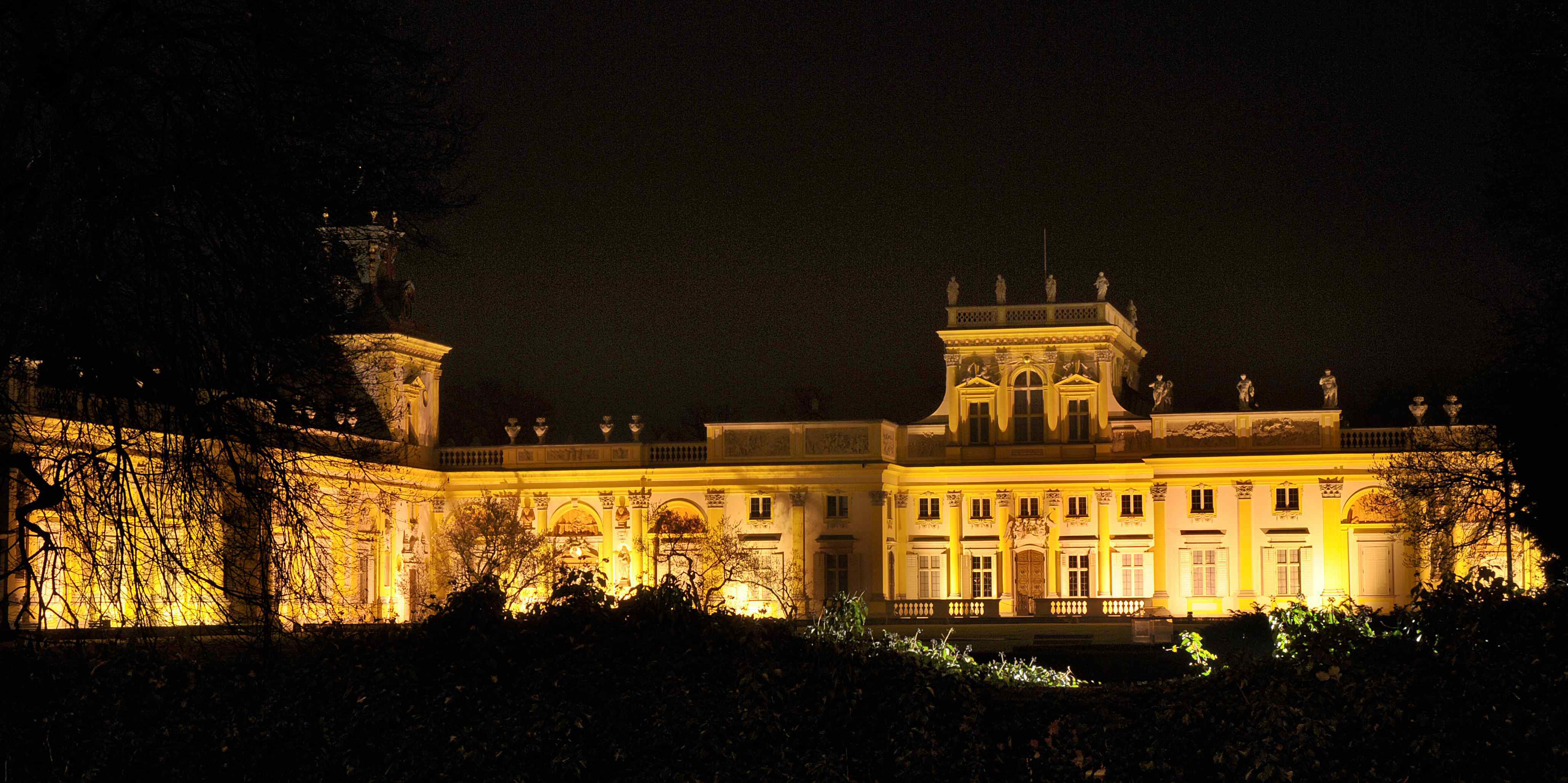 Kolorowe zdjęcie współczesne. Przedstawia oświetlony światłem Pałac w Wilanowie w nocy. Fasada jest złoto-żółta.