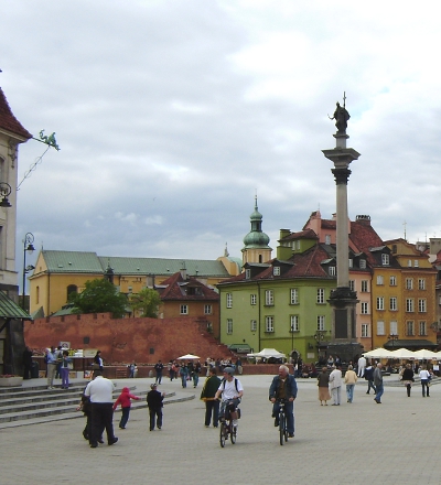 Kolorowe zdjęcie współczesne. Przedstawia widok na Starówkę warszawską. Na pierwszym planie spacerujący ludzie. W oddali kolumna Zygmunta, kamienice i fragment muru. Jest to widok na Plac  Zamkowy w Warszawie.