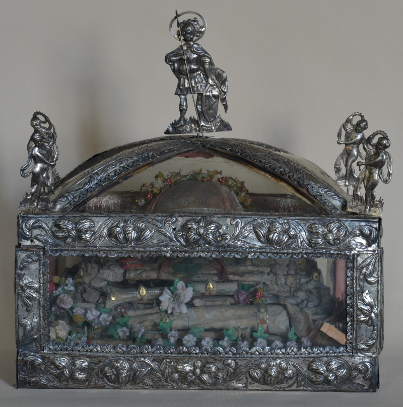 Przedmiot do przechowywania relikwii. Wykonany ze srebra i szkła. Jest to Relikwiarz św. Fortunata. Na szczycie postać świętego z tarczą i ostrzem. Po bokach tańczące postaci.