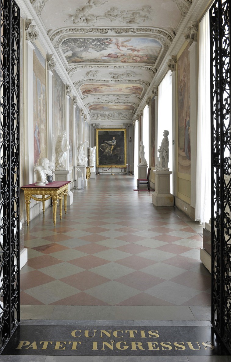 Kolorowe zdjęcie współczesne przedstawia korytarz palacowy na którym stoją rzeźby i meble antyczne. 