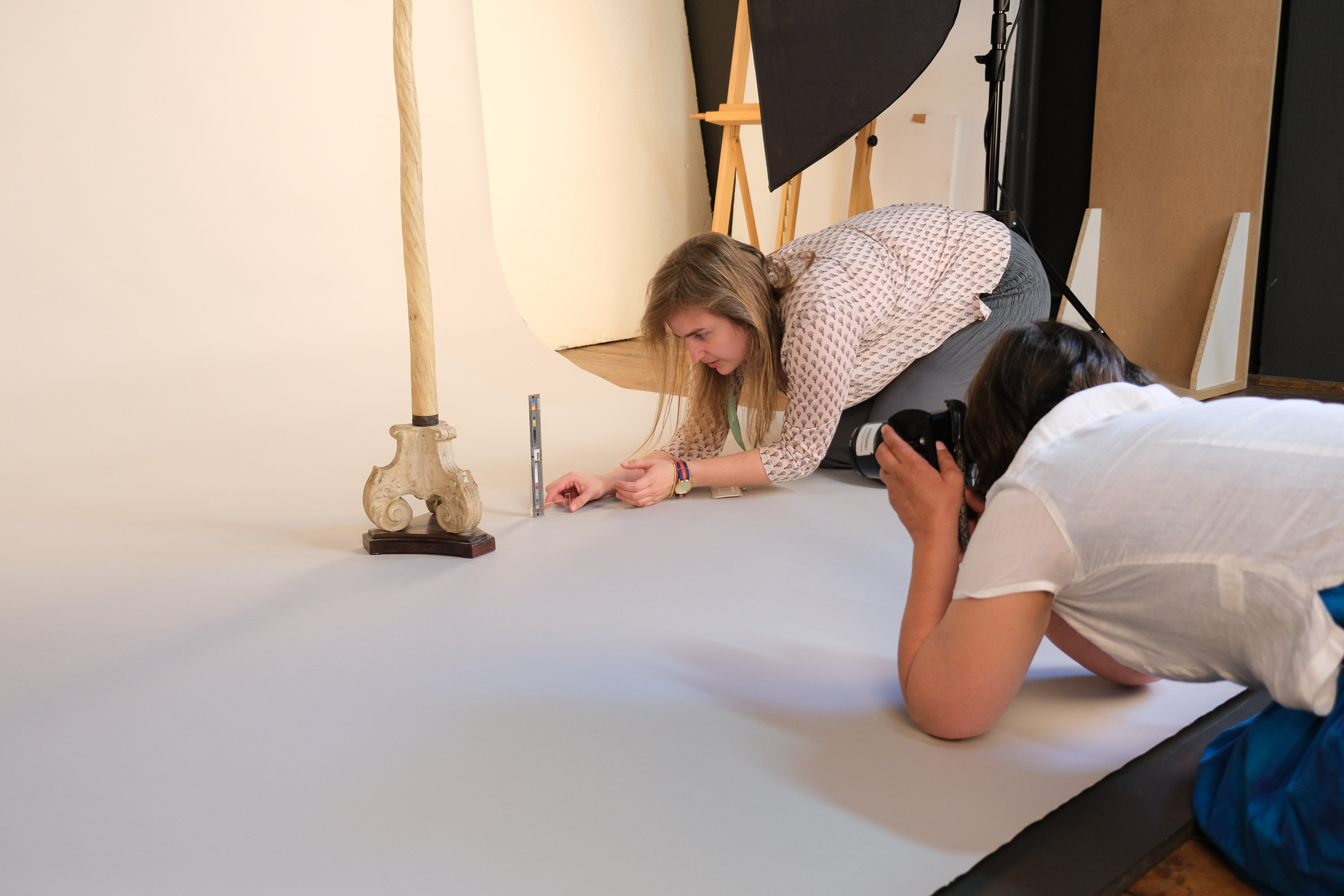 Zdjęcie pokazuje sesję fotograficzną. Na białym kartonie obiekt i dwie kobiety, które go fotografują. 