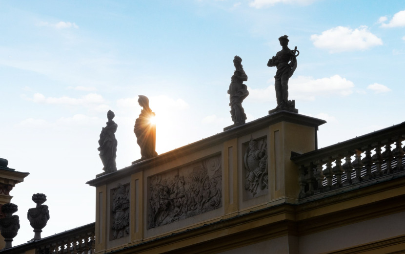 Współczesna kolorowa fotografia. Przedstawia widok na fragment szczytu pałacu w Wilanowie. Przy balustradzie podwyższenie na którym znajdują się cztery rzeźby. Zza rzeźb przebijają się promienie słońca.