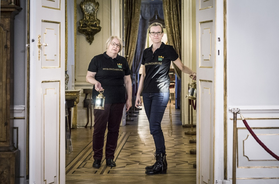 Contemporary colour photo. It depicts two middle-aged women dressed in black trousers and black short-sleeved shirts. They stand in the interior of the historic Palace. Before them is an open, white door.