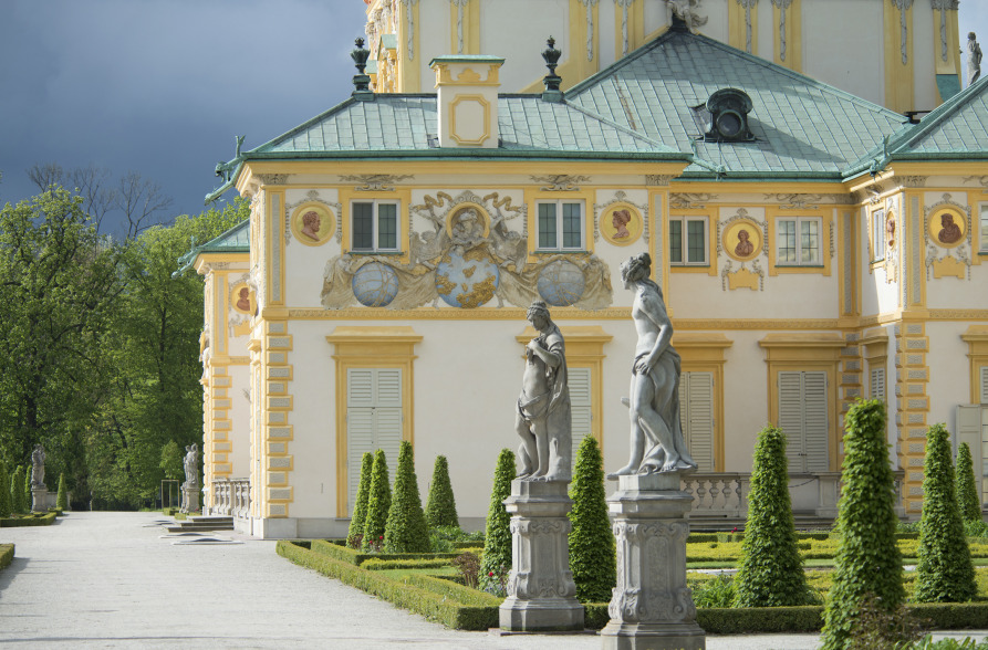 Kolorowe zdjęcie współczesne. Przedstawia widok na fragment niskiej budowli z wieżą, w kolorze jasnej żółci. Jest to Pałac w Wilanowie. Na pierwszym planie zielona trawa, drzewo i białe ławki.