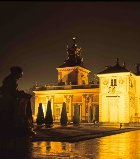 Współczesna kolorowa fotografia. Przedstawia fragment zabudowań pałacu w Wilanowie. Widzimy oświetloną na żółto architekturę.