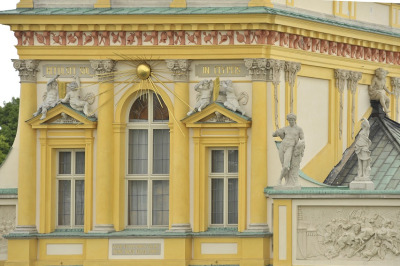 Fotografia kolorowa. Przedstawia ujęcie na fragment fasady budynku, gdzie widać trzy okna. Nad nimi płaskorzeźby i słońce w złocie. POwyżej dekoracyjny gzyms. 