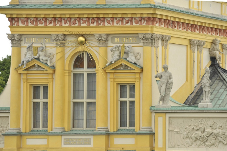 Fotografia kolorowa. Przedstawia ujęcie na fragment fasady budynku, gdzie widać trzy okna. Nad nimi płaskorzeźby i słońce w złocie. POwyżej dekoracyjny gzyms. 