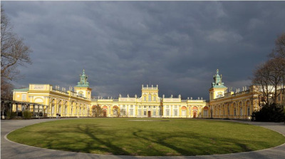 
Współczesna, kolorowa fotografia. Przedstawia pałac w Wilanowie oświetlony mocnym słońcem. Z przodu podjazd. Niebo ciemne, zachmurzone. Po bokach drzewa.
