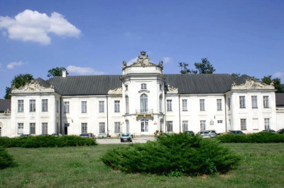 Zdjęcie kolorowe. Przedstawia pałac na tle pejzażu. Budynek jest biały, ma ciemno szary dach. 