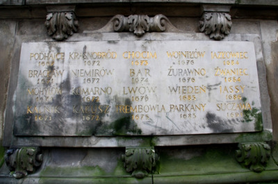 Fotografia kolorowa. Zbliżenie na pomnik konny króla Jana III w Gdańsku. Przedstawia zbliżenie na fragment płaskorzeźby na której widniej napisy miast i daty.