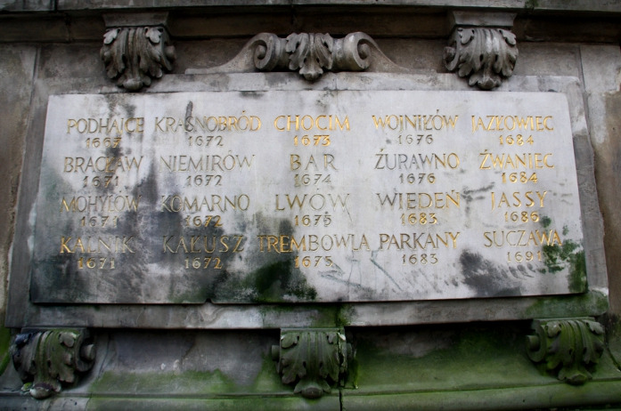 Fotografia kolorowa. Zbliżenie na pomnik konny króla Jana III w Gdańsku. Przedstawia zbliżenie na fragment płaskorzeźby na której widniej napisy miast i daty.