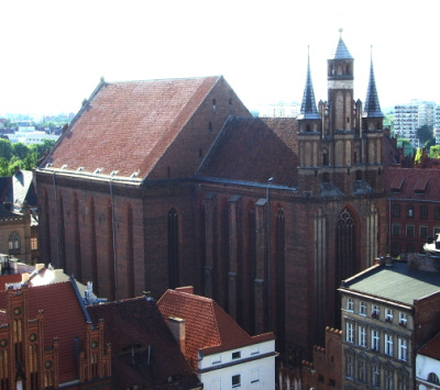 Fotografia kolorowa. Przedstawia gotycką bazylikę pośród innych zabudowań miejskich. Jest to kościół franciszkanów w Toruniu. 