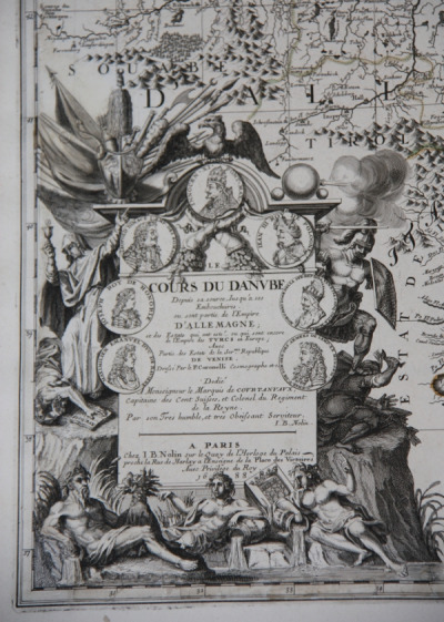 Fragment starej mapy. Przedstawia postaci mitologiczne. Na skale osadzona jest duża plansza papieru z medalionami i tekst.
