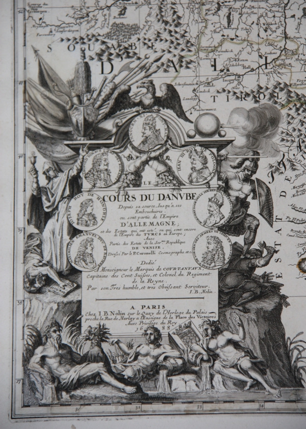Fragment starej mapy. Przedstawia postaci mitologiczne. Na skale osadzona jest duża plansza papieru z medalionami i tekst.