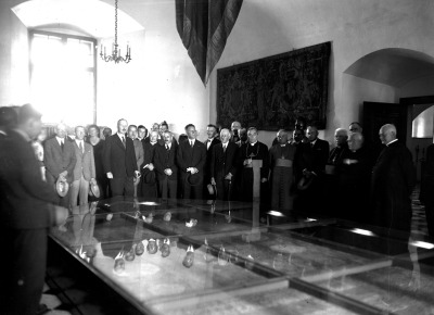 Archiwalne zdjęcie czarno-białe. Przedstawia zgromadzonych ludzi wokół stołu - poziomej gabloty w starej komnacie. Na jednej ze ścian wisi tkanina. Jest to wystawa pamiątek z epoki króla Jana III Sobieskiego na Zamku Królewskim na Wawelu.