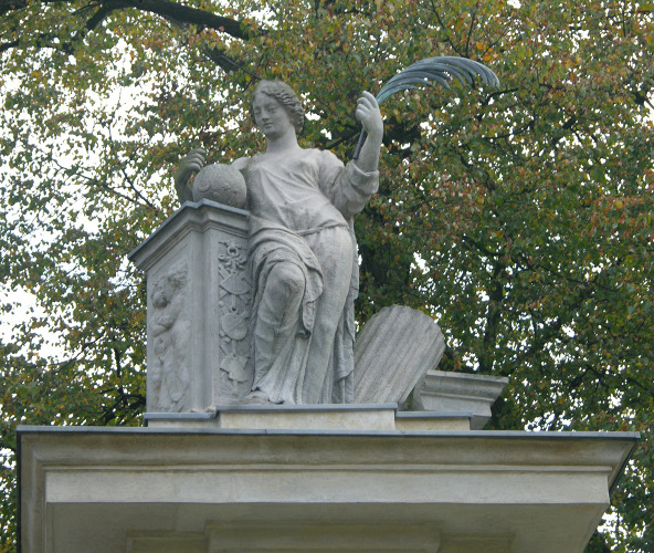 Fotografia kolorowa. Przedstawia rzeźbę - postać kobiety stojącej w kontrapoście. Kobieta ubrana w szaty antyczne opiera się o cokół na którym znajduje się kula ziemska. Kobieta trzyma w lewej dłoni gałązkę palmy. Obok niej leży fragment kolumny oparty o gzyms. Całość umieszczona na podniesieniu. Fotografia zrobiona z perspektywy żabiej. 