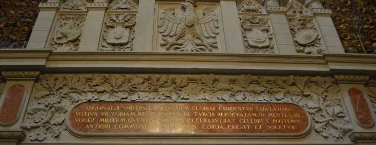 Zdjęcie kolorowe które przedstawia tablicę ze złotymi napisami, a wokół fragment dekoracji architektonicznej z motywami liści. Powyżej orzeł biały w koronie, a obok niego cztery herby – po prawej i lewej stronie. Dekoracja ta znajduje się na Wawelu. 