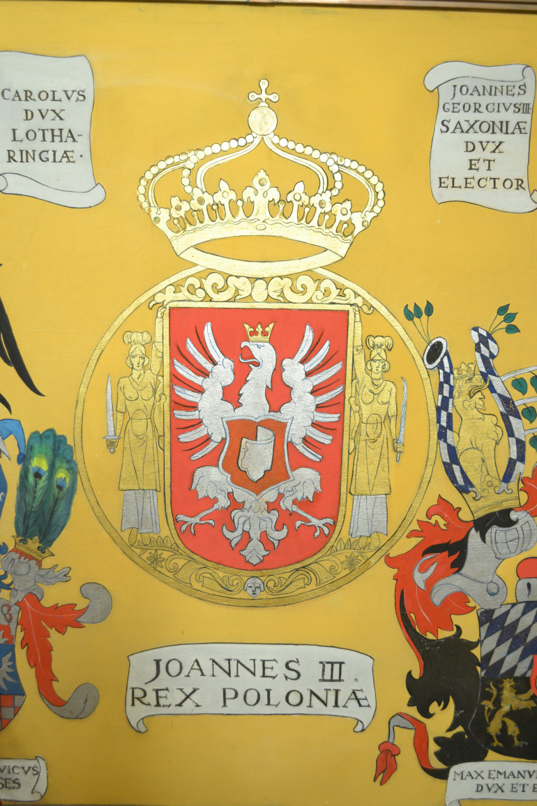 Zdjęcie przedstawia fragment fresu. Jest to herb Polski z orłem białym w koronie podtrzymywany prez dwie kobiety w szatach antycznych z mieczami. Nad godłem złota korona oparta o złotą obręcz. POd godłem napis: Joannes III REX POLONIAE. 