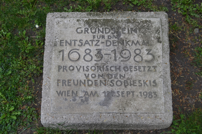 Zdjęcie przedstawia płytę kamienną osadzoną na trawniku. Jest to kamień węgielny pod pomnik Odsieczy Wiedeńskiej, który miał powstać w Wiedniu.  