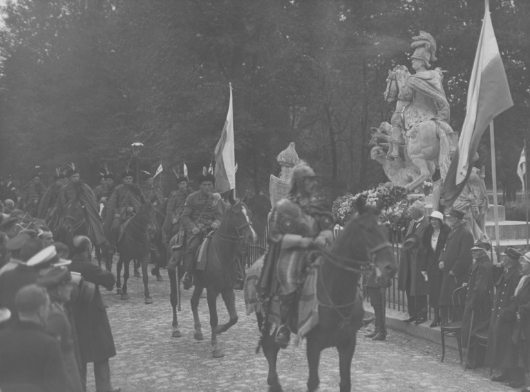 Archiwalne zdjęcie czarno-białe. Przedstawia żołnierzy na koniach, którzy jadą po alei obok której stoi pomnika króla Jana III Sobieskiego. Jest to dokumentacja z rocznicy odsieczy wiedeńskiej - uroczystości w Warszawie.