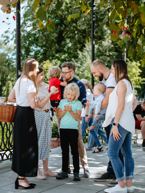 Kolorowe zdjęcie współczesne. Przedstawia grupę ludzi z dziećmi. Jest to spotkanie rodzin na tle pejzażu. Portretowani ubrani są w letnie stroje współczesne.