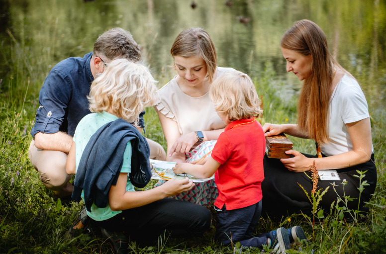 Colorful contemporary photo. A group of adults with children are sitting on the green grass. They are poring over a board game. Two little boys have their backs turned. The woman on the right holds a wooden box on her lap.