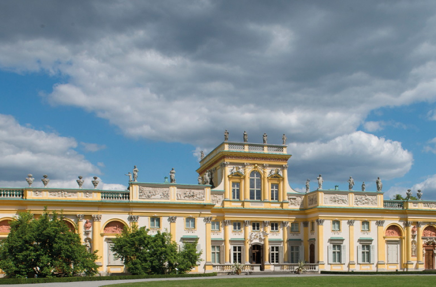 Colourful contemporary photo. It shows a view of a horizontal, low building with two towers, light yellow in colour. This is the Palace in Wilanów. 