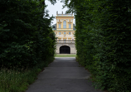 Widok na fragment budynku pałacu od strony korytarza utworzonego z wysokich krzewów.