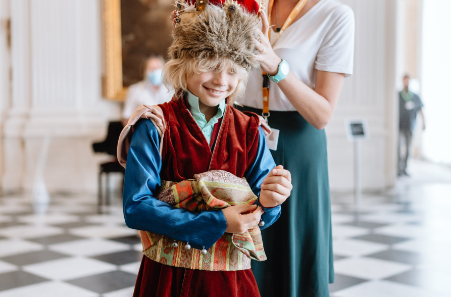 Młoda kobieta ubiera dziecko w strój historyczny. Chłopiec uśmiecha się. W głębi ludzie zwiedzający ekspozycję.