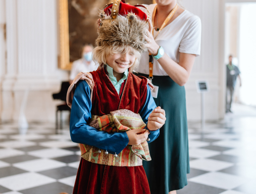 Młoda kobieta ubiera dziecko w strój historyczny. Chłopiec uśmiecha się. W głębi ludzie zwiedzający ekspozycję.