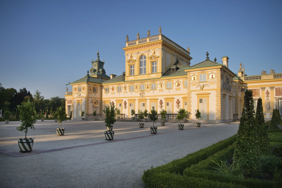 Widok na pałac w Wilanowie, który jest oświetlony słońcem. Na pierwszym planie plac, alejki i donice z drzewkami. 