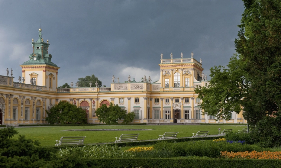 Widok na fragment niskiej budowli z wieżą, w kolorze jasnej żółci. Jest to Pałac w Wilanowie. Na pierwszym planie zielona trawa, drzewo i białe ławki.