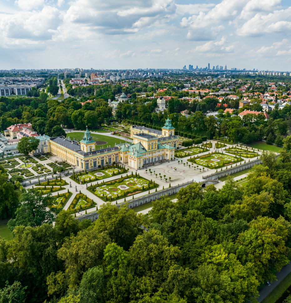 Widok z lotu ptaka na Pałac w Wilanowie otoczony ogrodami. W tle miasto Warszawa i nowoczesne blokowiska oraz dzienica domków jednorodzinnych. 