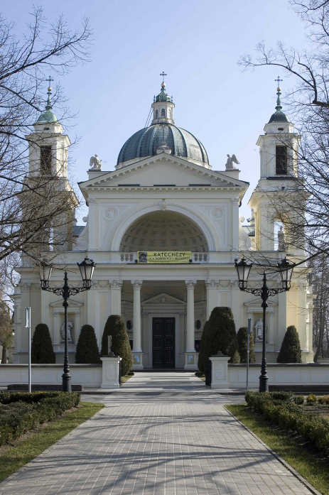 Kolorowa fotografia współczesna. Przedstawia biały kościół z kopułą i dwiema bocznymi wieżami. Przed kościołem ogrodzenie i deptak. Jest to Kościół św. Anny w Wilanowie.