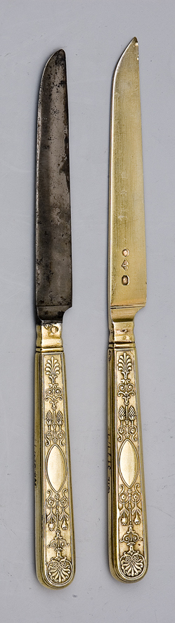 Zdjęcie przedstawia dwa złote noże deserowe, które mają ozdobne rączki. Powierzchnia rączek zdobiona motywami roślinnymi.