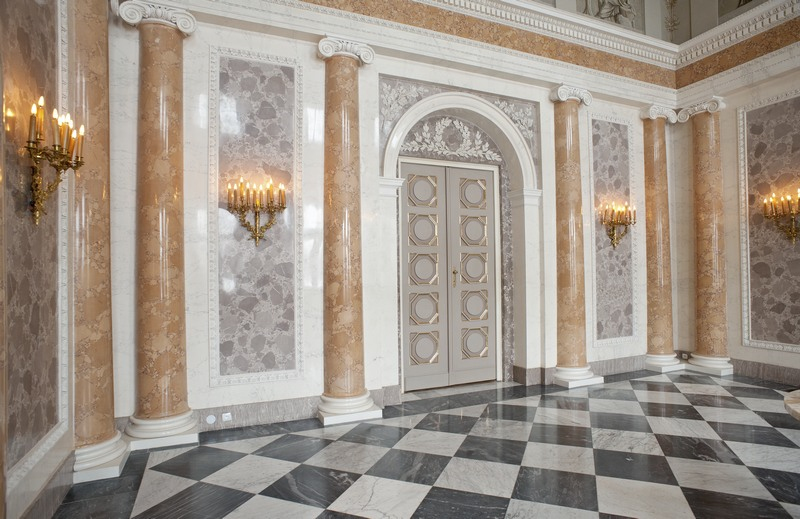 Kolorowe zdjęcie. Przedstawia salę z kolumnadą w pałacu. Na ścianach znajdują się złote świeczniki. Posadzka kamienna, dwukolorowa.