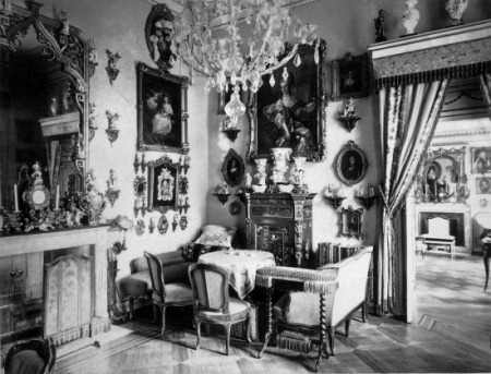 Czarno biała fotografia archiwalna. Przedstawia wnętrze pokoju w pałacu pełne antycznych mebli, obrazów i bibelotów. Jest to wnętrze Pokoju Saskiej Porcelany z widokiem na Gabinet Pastelowy (dawną sypialnię marszałkowej Lubomirskiej).