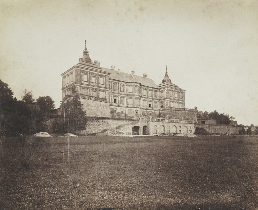Stara, czarno-biała fotografia archiwalna. Przedstawia duży zamek na tle pejzażu. Przed zamkiem duża połać trawy. Jest to zamek w Podhorcach. 