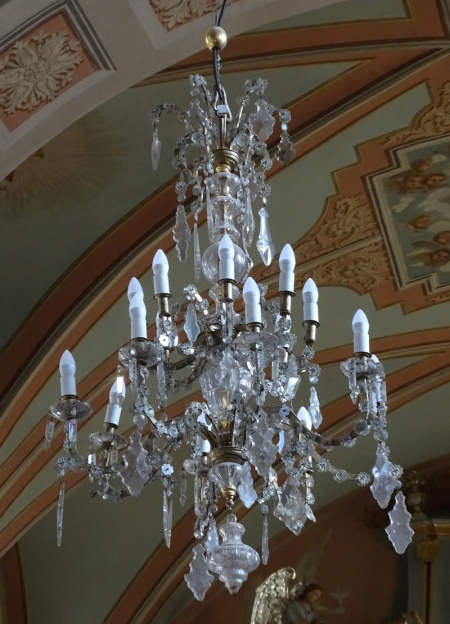 Kolorowe zdjęcie przedstawia żyrandol kryształowy, umieszczony tuż pod sklepieniem. Żyrandol ma białe, wysmukłe żarówki.