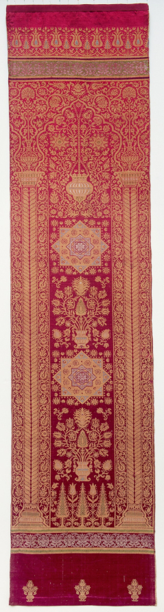 Zdjęcie przedstawia zabytkową tkaninę. Jest to makata typu arkadowego wykonana w Turcji. Uszyta z atłasu wzorzystego i jedwabiu. Zdobienia motywami roślinnymi przy użyciu nici z metalowym oplotem. 