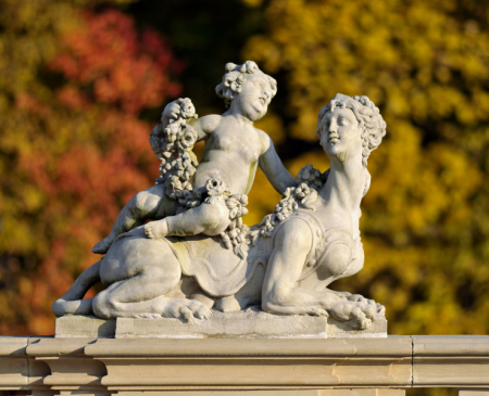 Kolorowe zdjęcie współczesne. Przedstawia rzeźbę na balustradzie. Jest to putto ze sfinksem. Putto siedzi na grzbiecie sfinksa. Trzyma w rękach girlandę z kwiatami. Jest na to rzeźba z warsztatu J.Ch. Redlera.