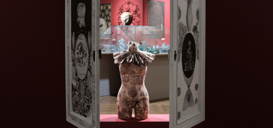 Manekin kobiecy ozdobiony kryzą i graficznymi wzorami stojący pomiędzy uchylonymi drzwiami.