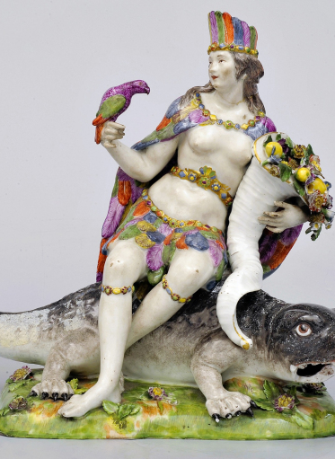 Porcelanowa figurka kobiety ubranej w kolorowe pióra siedzącej na krokodylu, trzymającej papugę w jednej dłoni oraz róg obfitości