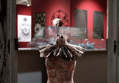 Ozdobiony kryzą i czarnobiałymi motywami kobiecy manekin na tle czerwonej ściany z obrazami