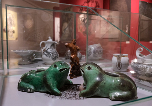 Dwa naczynia w kształcie zielonych żab, drewniana figurka postaci ludzkiej, naczynia w czarne wzory.