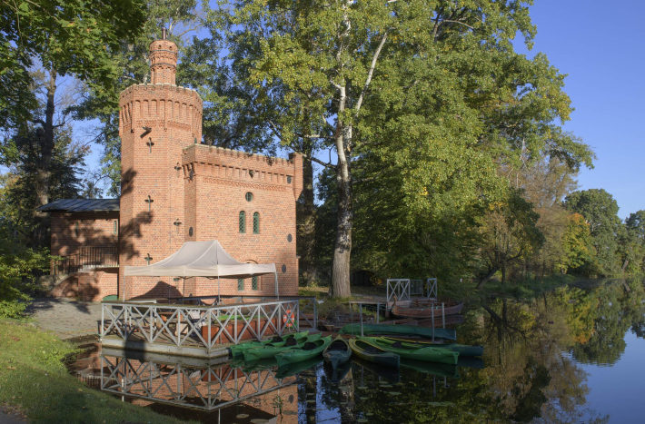 Budynek pompowni przypominający ceglany niewielki zamek otoczony drzewami. Przed nim Jezioro Wilanowskie i kajaki.